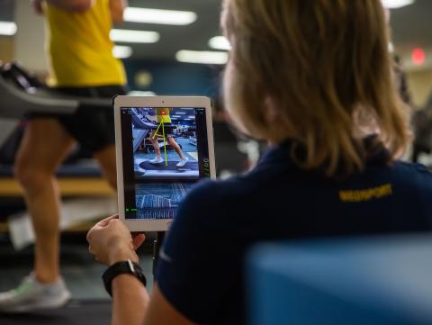 Woman records a video of a person running on a treadmill in the background.