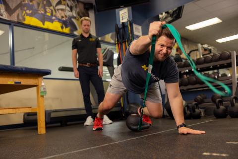 Man makes a move using a rope and a kettlebell inside a gym.