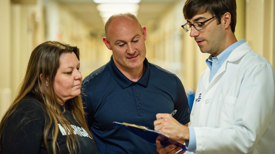 Dr. Jakob McSparron wearing white coat and writing on clipboard talking to bald man in blue polo shirt and woman with long brown hair wearing blue t-shirt with word Michigan in white letters