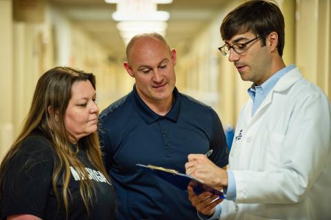 Dr. Jakob McSparron wearing white coat and writing on clipboard talking to bald man in blue polo shirt and woman with long brown hair wearing blue t-shirt with word Michigan in white letters